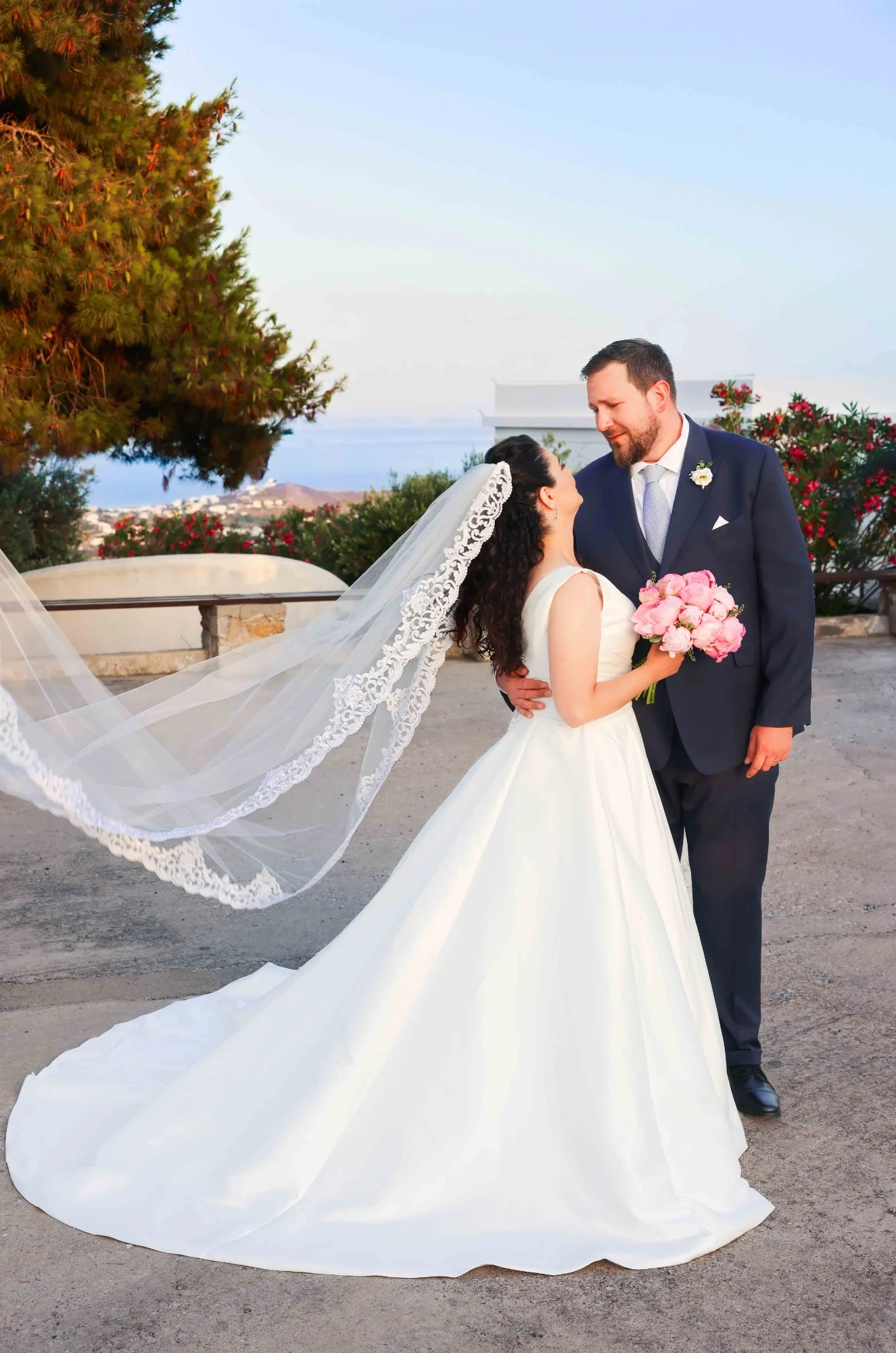 Bride and groom standing outdoors, bride in a white dress holding pink flowers, veil flowing behind, groom in a dark suit. Gabriel Lenca Photography - London Wedding Photographer.