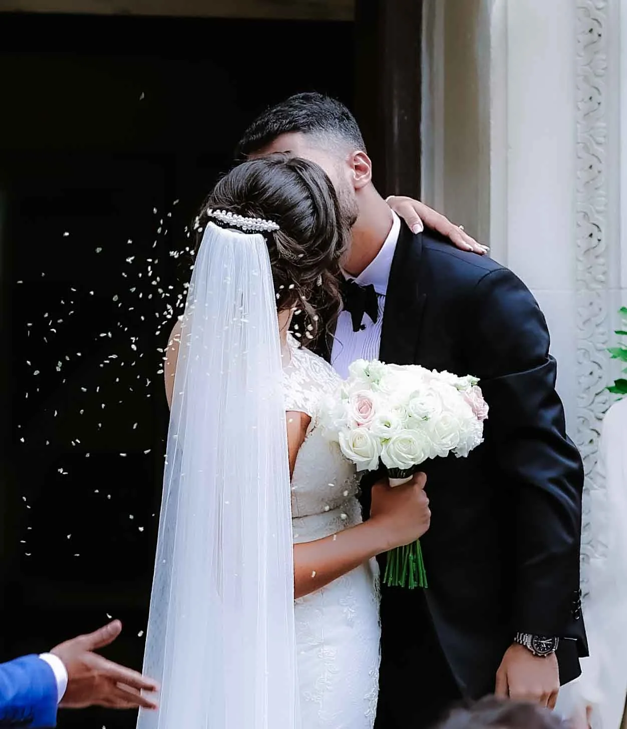A bride and groom kiss at their wedding. The bride holds a bouquet of white roses and wears a long veil. The groom is in a black suit. Rice or confetti is tossed in the background. Gabriel Lenca Photography - London Wedding Photographer.