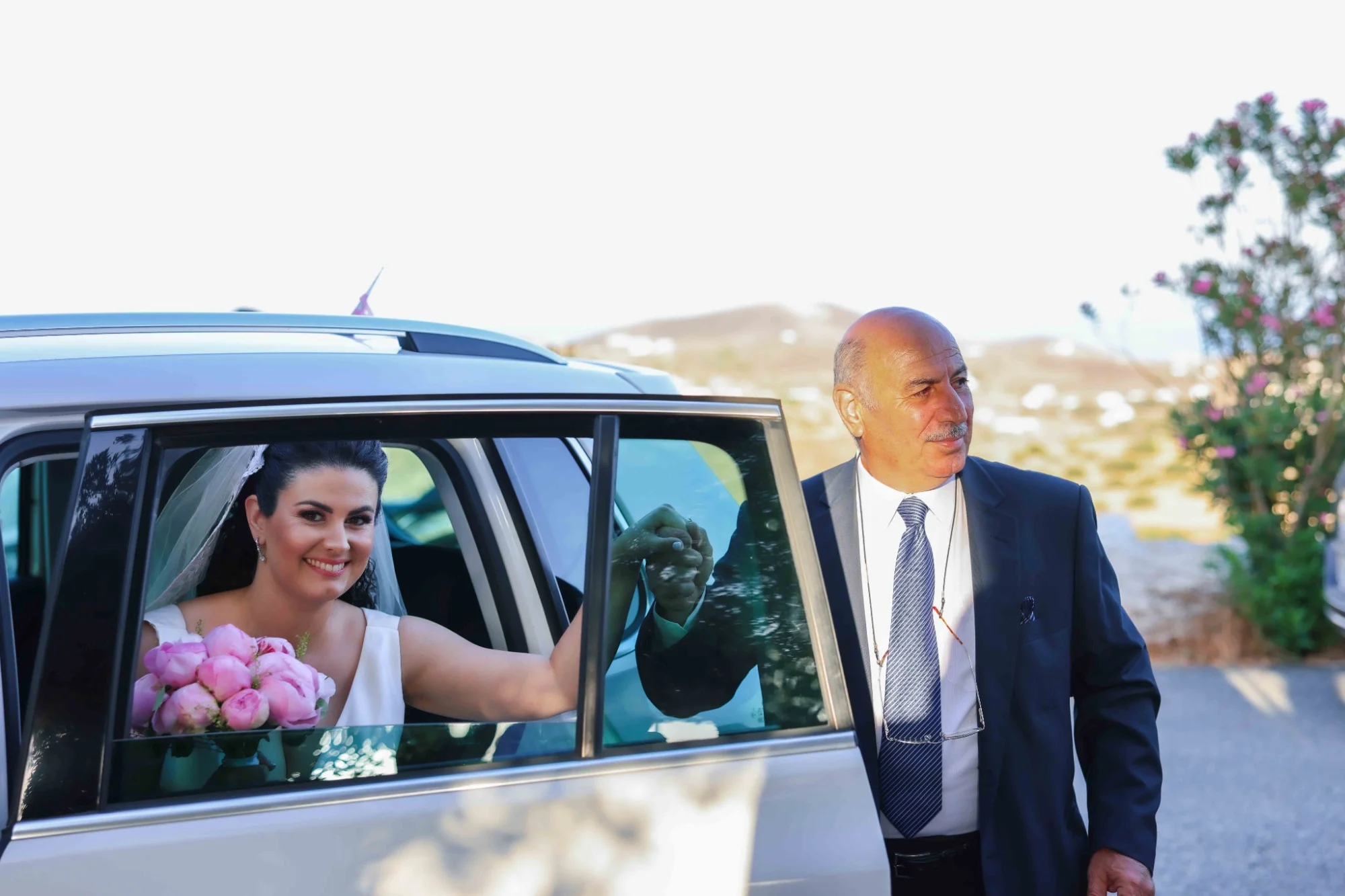 A smiling bride holding pink flowers sits in a car, while a man in a suit stands outside, holding her hand through the window. Gabriel Lenca Photography - London Wedding Photographer.