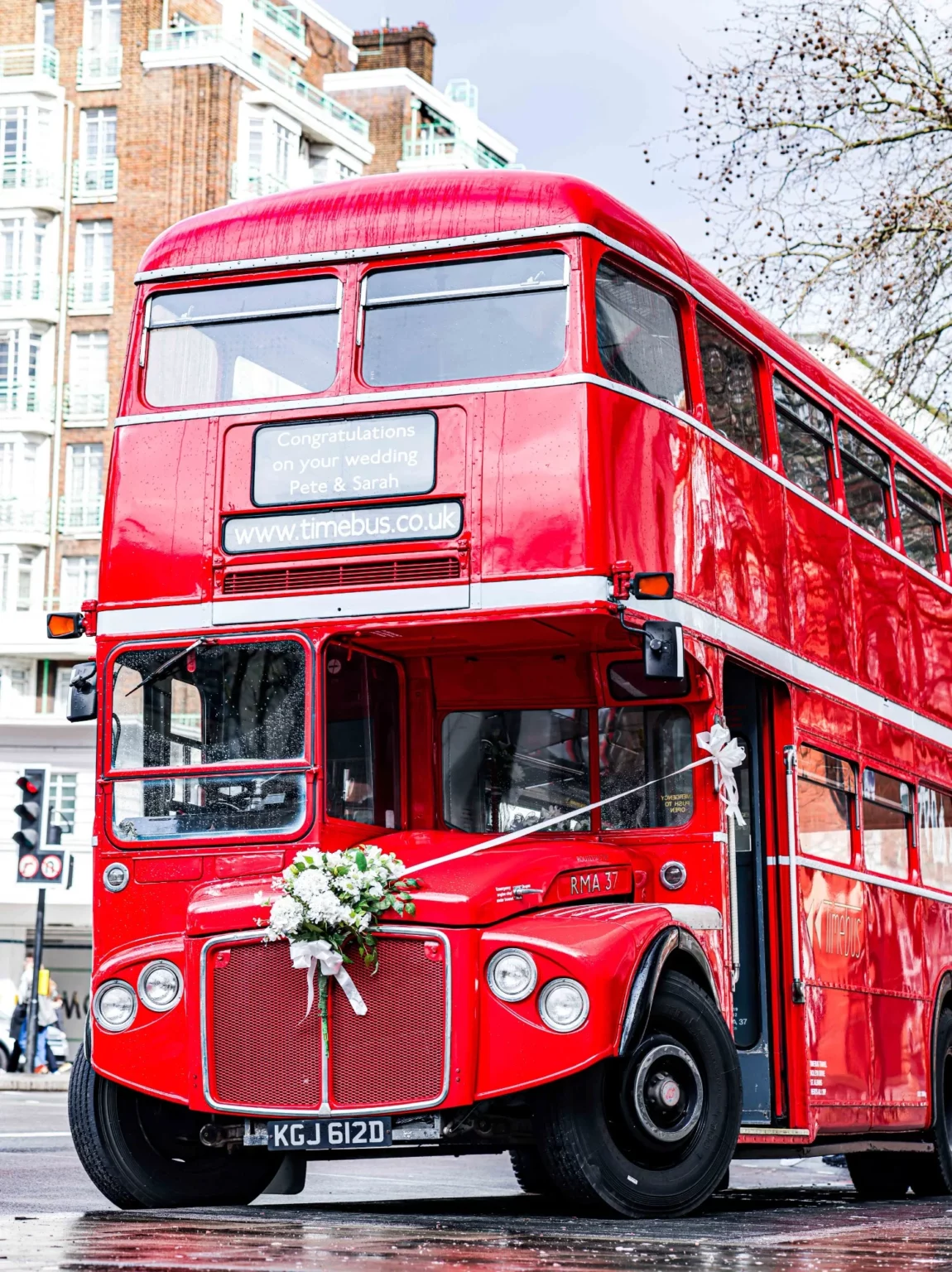 A red double-decker bus with wedding decorations, including white flowers on the front, parked on a wet city street. Gabriel Lenca Photography - London Wedding Photographer.