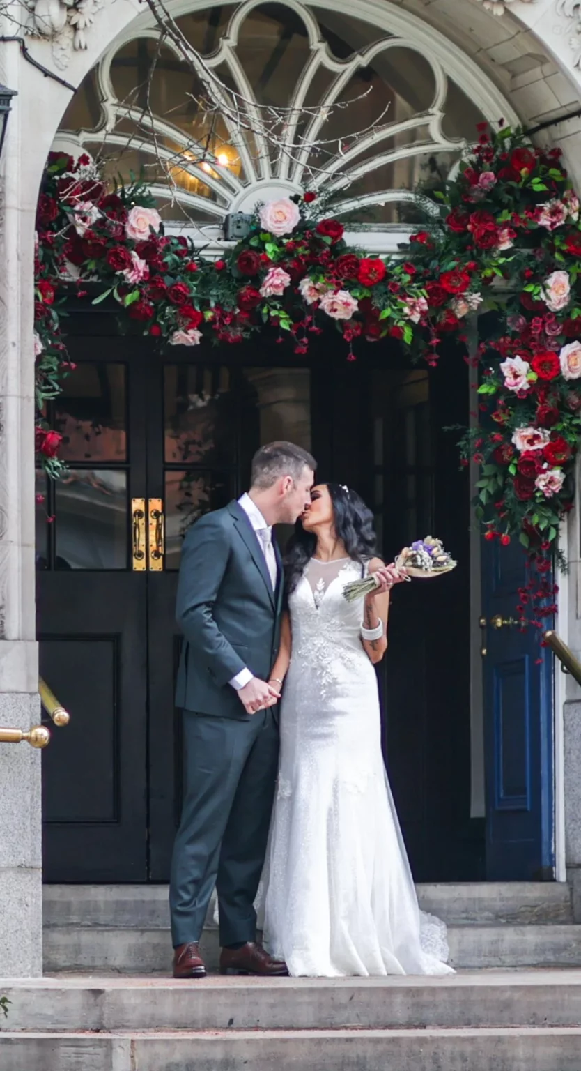 Couple in wedding attire shares a kiss under a floral archway outside a building. The bride holds a bouquet. Gabriel Lenca Photography - London Wedding Photographer.