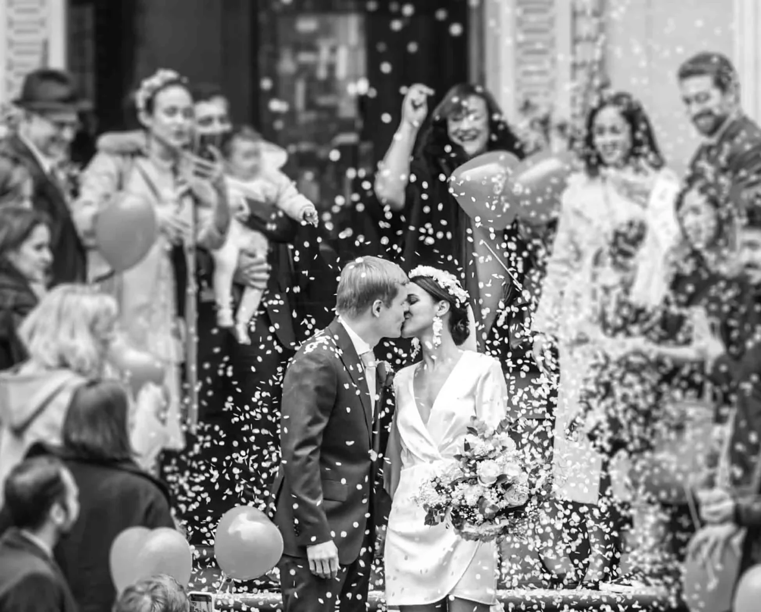 A couple, dressed in wedding attire, kisses amidst a crowd throwing confetti and holding balloons outside a building. Gabriel Lenca Photography - London Wedding Photographer.