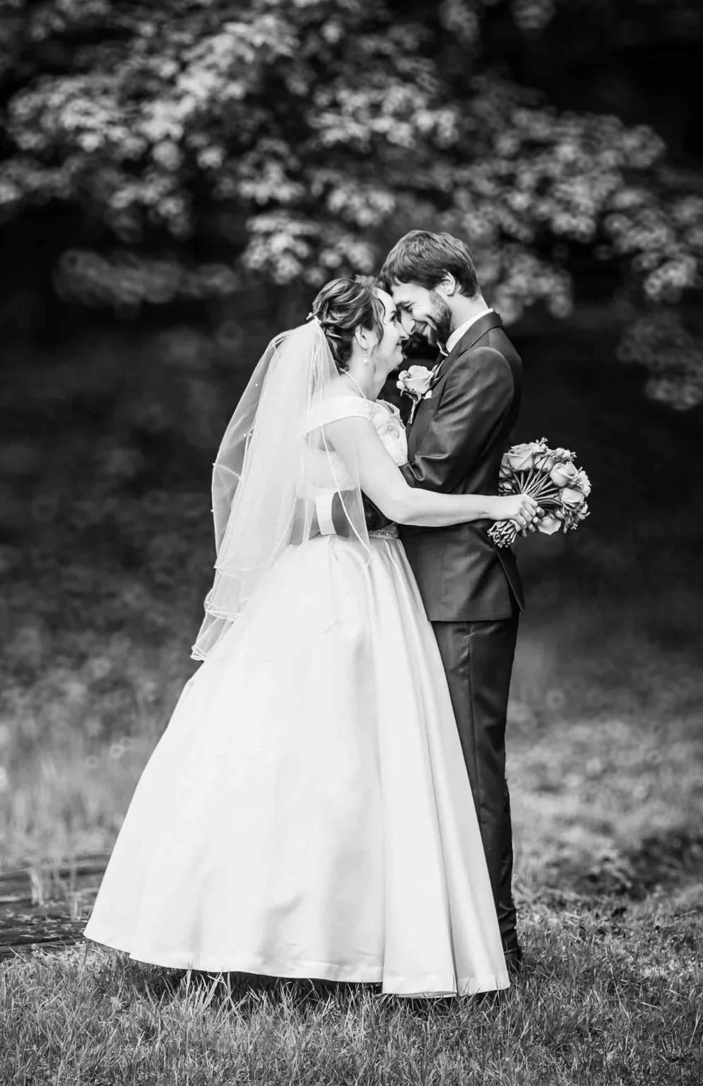 A bride and groom share an intimate moment outdoors. The bride holds a bouquet and wears a veil and gown, while the groom wears a suit. The background is softly blurred foliage. Gabriel Lenca Photography - London Wedding Photographer.