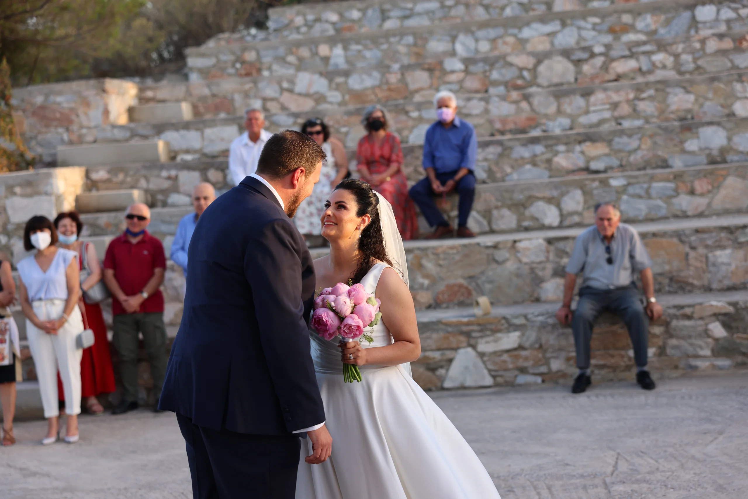 Bride and groom smiling outdoors with guests seated.